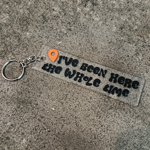 May include: A rectangular clear acrylic keyring featuring a silver metal split ring and a small orange map pin icon. The text "I've been here the whole time" is printed in black, bold, rounded lettering across the surface of the transparent tag.
