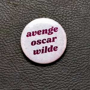 Puede incluir: Botón blanco con el texto "avenge oscar wilde" en letras moradas.