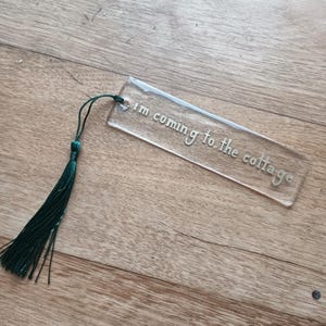 Könnte beinhalten: Ein klarer Acryl-Lesezeichen mit dem Schriftzug "I'm coming to the cottage" in kursiver Schrift. Am oberen Ende des Lesezeichens ist eine dunkelgrüne Quaste befestigt. Das Lesezeichen ist rechteckig.