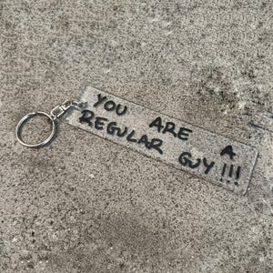 Puede incluir: Un llavero rectangular de acrílico transparente con una anilla de metal plateado y un cierre de mosquetón. El llavero tiene el texto negro escrito a mano "YOU ARE A REGULAR GUY !!!" impreso en la superficie con una fuente sans-serif.