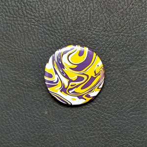 May include: A round button with a swirling pattern in yellow, white, and purple.
