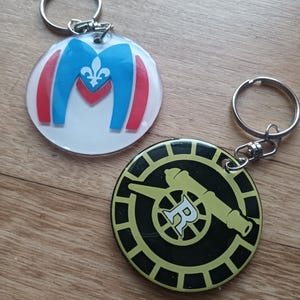 Puede incluir: Dos llaveros con aros plateados. Uno es redondo con fondo blanco y diseño azul, rojo y blanco. El otro es un círculo negro con un diseño de cañón amarillo y la letra "R".