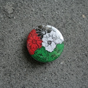Puede incluir: Un botón redondo con un diseño floral. El botón presenta un diseño segmentado con flores rojas, blancas, verdes y negras. El botón está sobre un fondo gris texturizado.