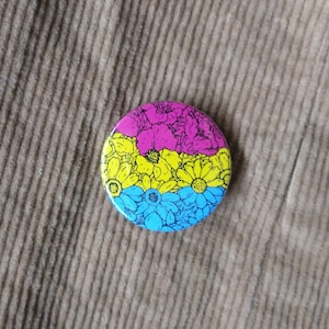 Può includere: Un bottone rotondo con un design floreale in blu, giallo e rosa. I fiori sono stilizzati e assomigliano a una bandiera arcobaleno.