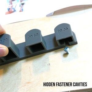 May include: A close-up of a gray plastic tool with four cavities labeled "20-35", "30-60", "40-80", and "50-100". The tool is used to create hidden fastener cavities in wood. A screw is visible in the background.