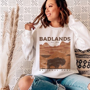 K&ouml;nnte beinhalten: Wei&szlig;er Pullover mit einem braunen Grafikdruck einer Landschaft und einem B&uuml;ffel. Der Grafiktext lautet "Badlands Est. 1978 National Park".