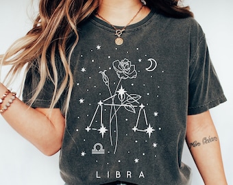 Op de afbeelding: Een donkergrijs T-shirt met een wit grafisch ontwerp van een roos en een sterrenbeeld met het woord "LIBRA" eronder. Het grafische ontwerp doet denken aan een sterrenbeeld.