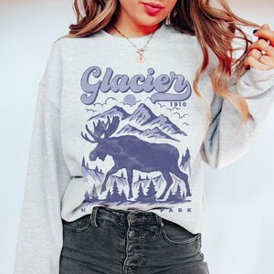 May include: A gray sweatshirt with a purple graphic design of a moose standing in front of mountains. The text "Glacier 1910" is above the moose and "National Park" is below the moose.