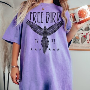 Puede incluir: Una camiseta de gran tama&ntilde;o de color morado con un gr&aacute;fico negro de un p&aacute;jaro en vuelo. El gr&aacute;fico incluye el texto "FREE BIRD" y los n&uacute;meros "19" y "73".