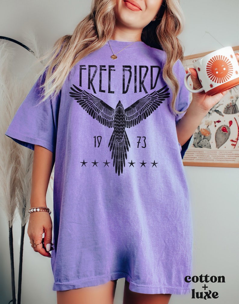 Puede incluir: Una camiseta de gran tama&ntilde;o de color morado con un gr&aacute;fico negro de un p&aacute;jaro en vuelo. El gr&aacute;fico incluye el texto "FREE BIRD" y los n&uacute;meros "19" y "73".