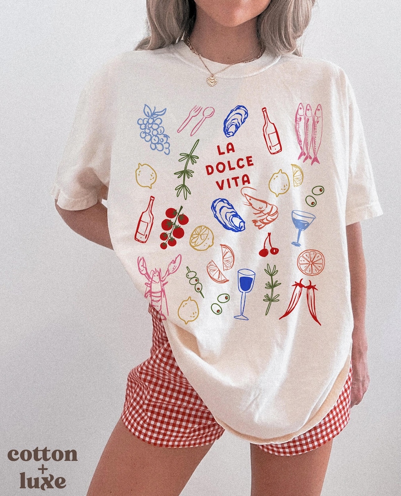 K&ouml;nnte beinhalten: Cremefarbenes T-Shirt mit einer farbenfrohen Illustration italienischer Speisen und Getr&auml;nke, darunter Trauben, Zitronen, Austern, Weinflaschen und dem Text "LA DOLCE VITA". Das Shirt wird mit rot-wei&szlig; karierten Shorts kombiniert.