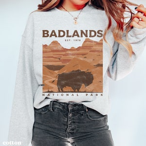 K&ouml;nnte beinhalten: Ein graues Sweatshirt mit einem braunen Aufdruck eines Bisons vor einer Landschaft. Der Aufdruck lautet "Badlands National Park Est. 1978".