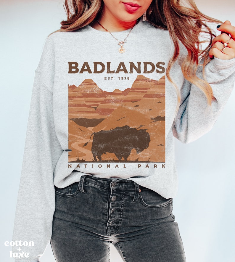 K&ouml;nnte beinhalten: Ein graues Sweatshirt mit einem braunen Aufdruck eines Bisons vor einer Landschaft. Der Aufdruck lautet "Badlands National Park Est. 1978".