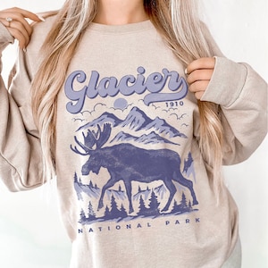 May include: A beige sweatshirt with a purple graphic design of a moose standing in front of mountains with the text "Glacier 1910 National Park".