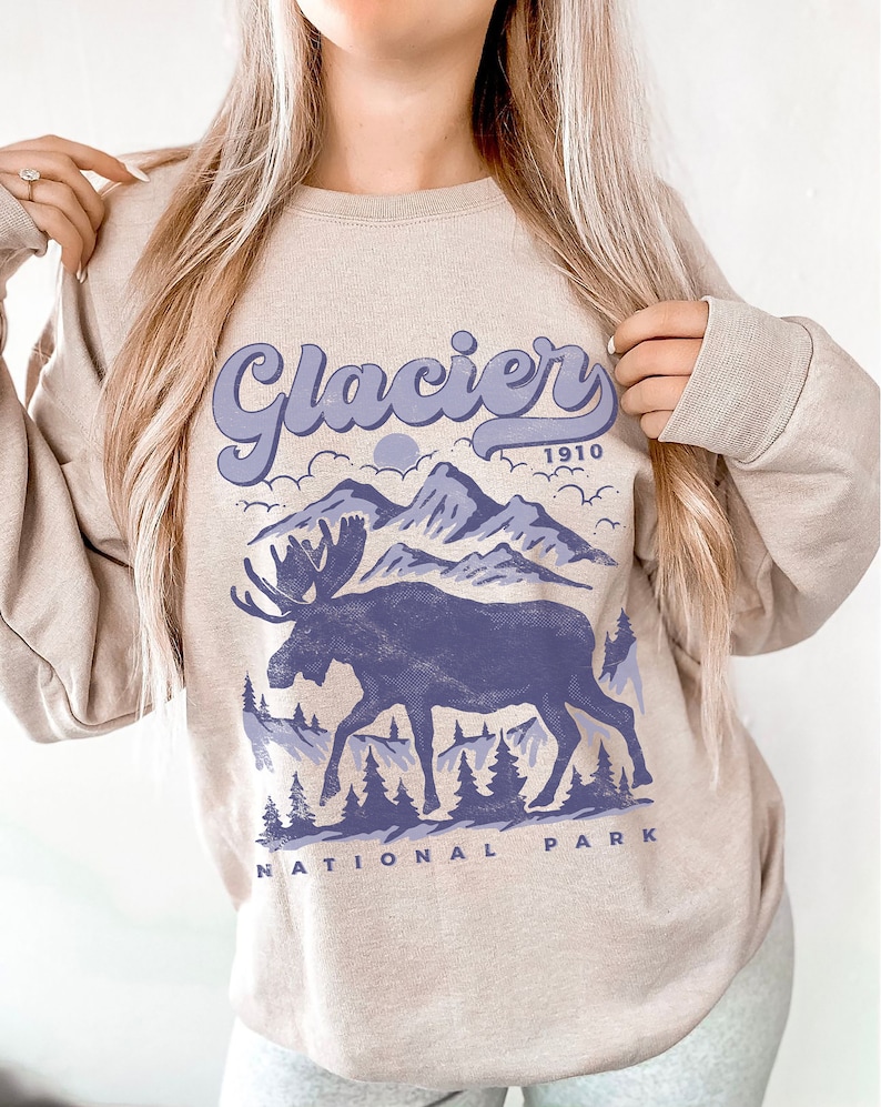 May include: A beige sweatshirt with a purple graphic design of a moose standing in front of mountains with the text "Glacier 1910 National Park".