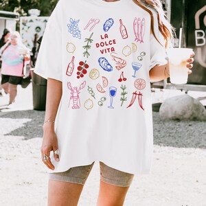 K&ouml;nnte beinhalten: Wei&szlig;es Baumwoll-T-Shirt mit einem farbenfrohen Grafikdesign mit Illustrationen von Speisen und Getr&auml;nken, darunter Trauben, Austern, Flaschen und ein Hummer. Der Text "LA DOLCE VITA" ist auf dem T-Shirt zentriert.