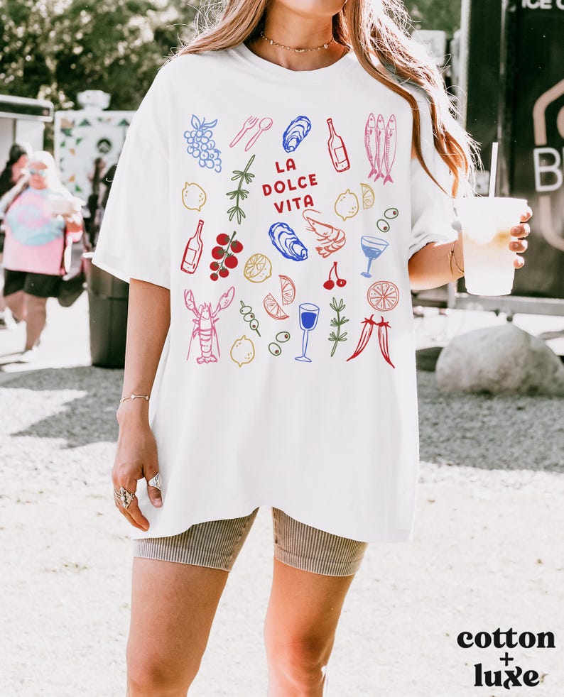 K&ouml;nnte beinhalten: Wei&szlig;es Baumwoll-T-Shirt mit einem farbenfrohen Grafikdesign mit Illustrationen von Speisen und Getr&auml;nken, darunter Trauben, Austern, Flaschen und ein Hummer. Der Text "LA DOLCE VITA" ist auf dem T-Shirt zentriert.