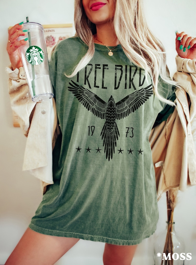 Puede incluir: Una camiseta verde con un gr&aacute;fico negro de un p&aacute;jaro con el texto "FREE BIRD" y los n&uacute;meros "19 73" en la parte delantera. La camiseta tiene un aspecto desgastado.