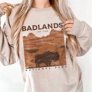 K&ouml;nnte beinhalten: Ein beiger Pullover mit einem Grafikdruck des Badlands National Park. Die Grafik zeigt eine braune Bisonsilhouette vor einer Landschaft aus roten und orangefarbenen H&uuml;geln. Der Text "Badlands National Park" und "Est. 1978" ist in Braun gedruckt.