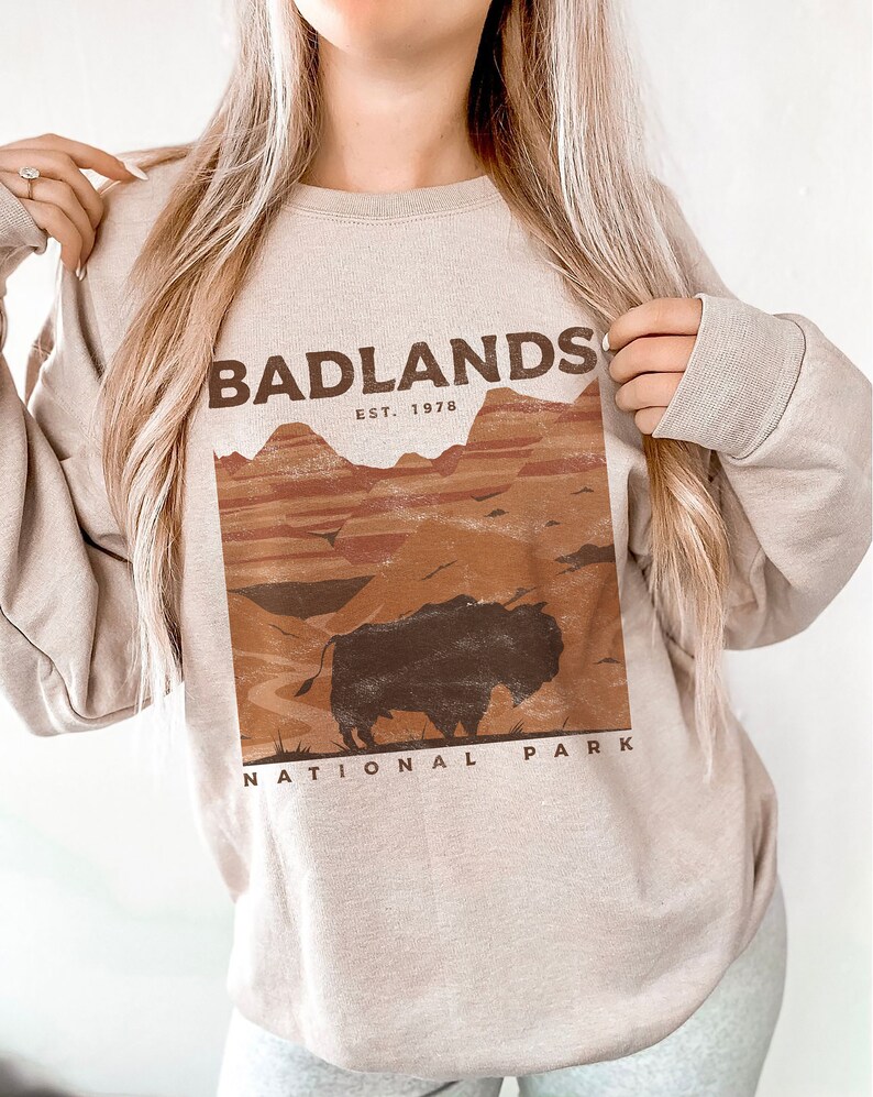 K&ouml;nnte beinhalten: Ein beiger Pullover mit einem Grafikdruck des Badlands National Park. Die Grafik zeigt eine braune Bisonsilhouette vor einer Landschaft aus roten und orangefarbenen H&uuml;geln. Der Text "Badlands National Park" und "Est. 1978" ist in Braun gedruckt.