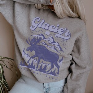 May include: A gray sweatshirt with a purple graphic of a moose standing in front of mountains with the text "Glacier National Park 1910" printed on it.