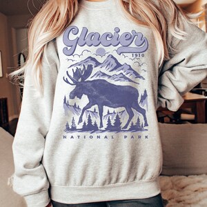 May include: A gray sweatshirt with a purple moose and mountain graphic. The text "Glacier 1910 National Park" is printed on the sweatshirt.