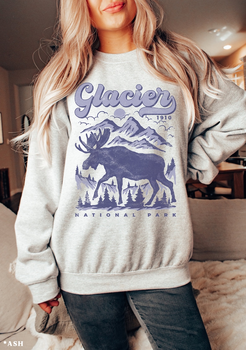 May include: A gray sweatshirt with a purple moose and mountain graphic. The text "Glacier 1910 National Park" is printed on the sweatshirt.