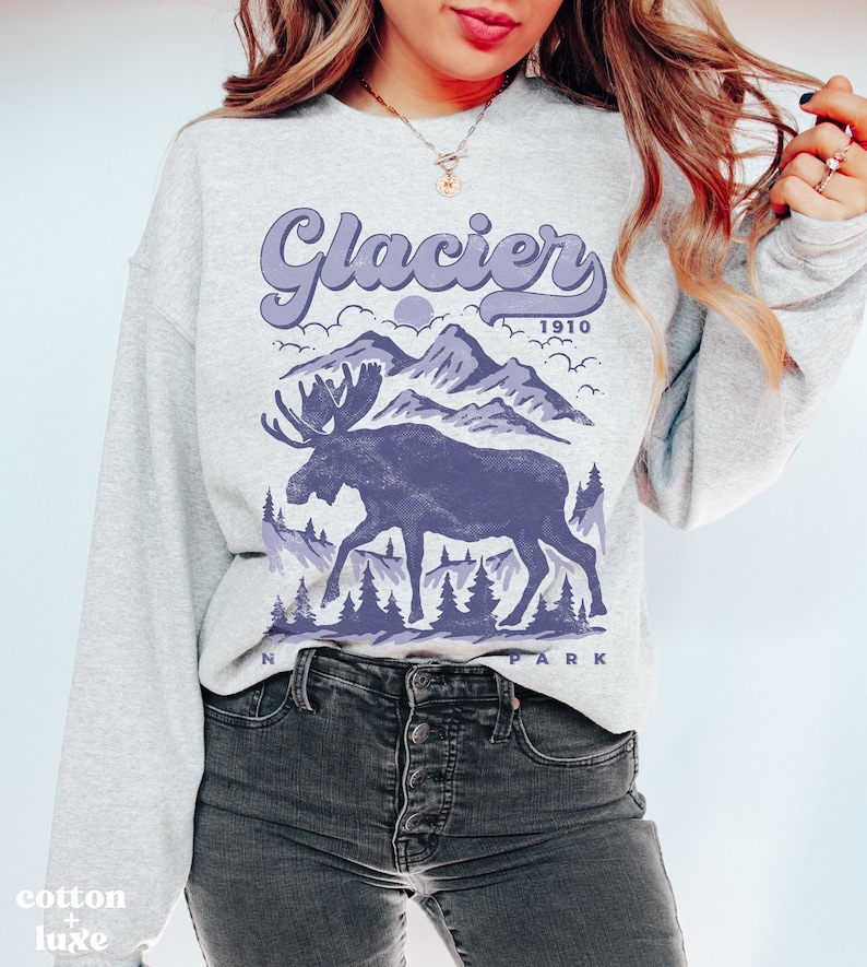 May include: A gray sweatshirt with a purple graphic design of a moose standing in front of mountains. The text "Glacier 1910" is above the moose and "National Park" is below the moose.