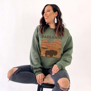 K&ouml;nnte beinhalten: Ein gr&uuml;nes Sweatshirt mit einem Grafikdruck eines Sonnenuntergangs und eines Bisons. Der Text "Badlands National Park Est. 1939" ist auf dem Sweatshirt aufgedruckt.