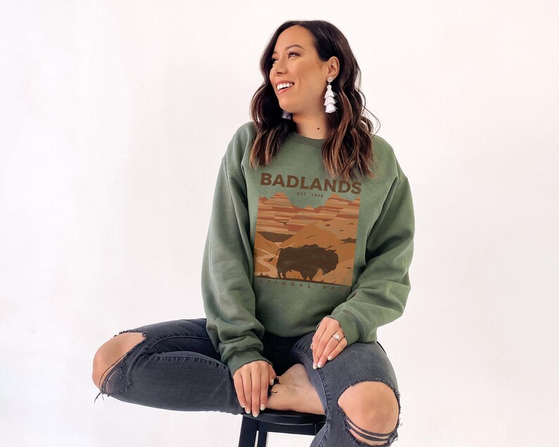 K&ouml;nnte beinhalten: Ein gr&uuml;nes Sweatshirt mit einem Grafikdruck eines Sonnenuntergangs und eines Bisons. Der Text "Badlands National Park Est. 1939" ist auf dem Sweatshirt aufgedruckt.
