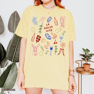 K&ouml;nnte beinhalten: Hellgelbes T-Shirt mit einem farbenfrohen Grafikdesign mit Illustrationen von Speisen und Getr&auml;nken, darunter Trauben, Austern, Flaschen und ein Hummer. Der Text "LA DOLCE VITA" befindet sich in der Mitte. Das Shirt ist aus Baumwolle.
