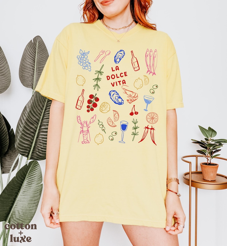 K&ouml;nnte beinhalten: Hellgelbes T-Shirt mit einem farbenfrohen Grafikdesign mit Illustrationen von Speisen und Getr&auml;nken, darunter Trauben, Austern, Flaschen und ein Hummer. Der Text "LA DOLCE VITA" befindet sich in der Mitte. Das Shirt ist aus Baumwolle.