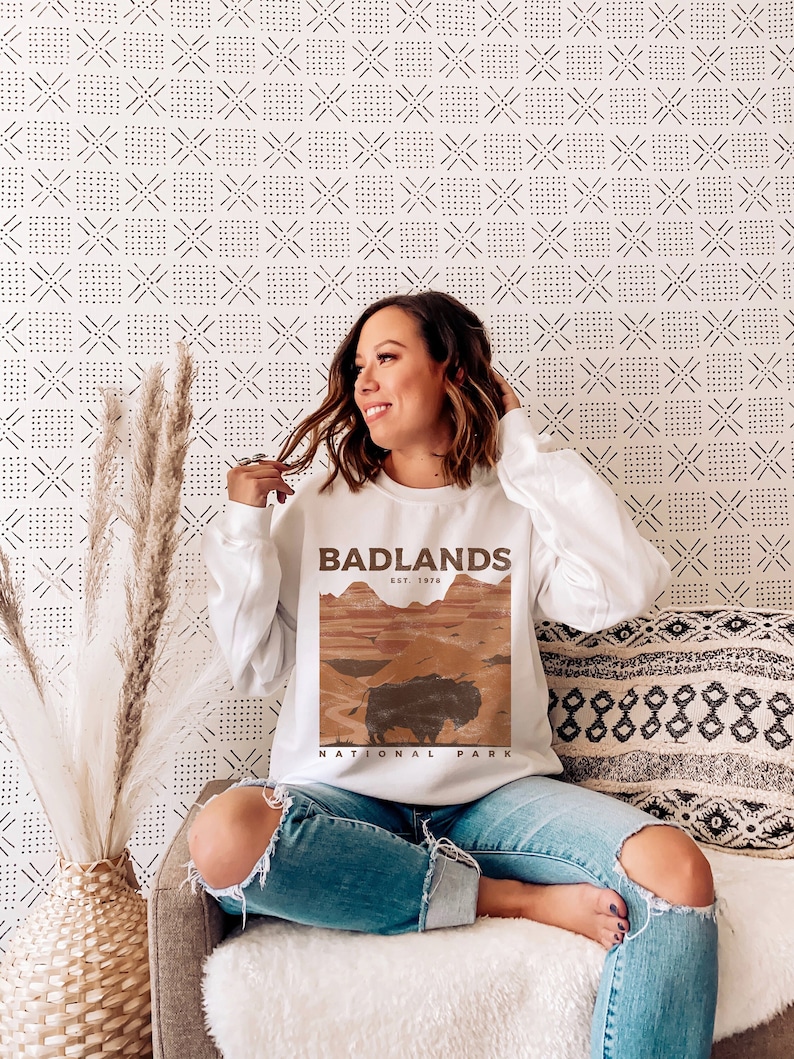 K&ouml;nnte beinhalten: Wei&szlig;er Pullover mit einem braunen Grafikdruck einer Landschaft und einem B&uuml;ffel. Der Grafiktext lautet "Badlands Est. 1978 National Park".