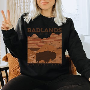 K&ouml;nnte beinhalten: Schwarzes Sweatshirt mit einem braunen Grafikdruck einer Landschaft mit einem Bison und dem Text "Badlands National Park Est. 1978."