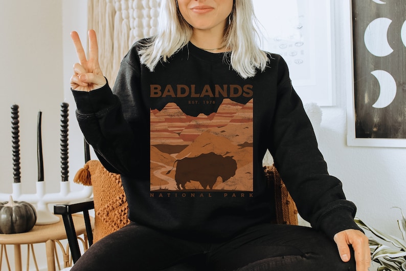 K&ouml;nnte beinhalten: Schwarzes Sweatshirt mit einem braunen Grafikdruck einer Landschaft mit einem Bison und dem Text "Badlands National Park Est. 1978."
