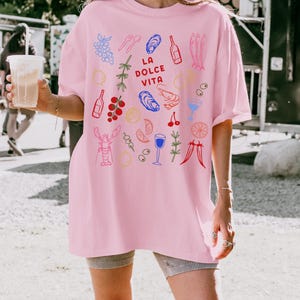 K&ouml;nnte beinhalten: Hellrosa Oversize-T-Shirt mit farbigen Illustrationen von Speisen und Getr&auml;nken, darunter Trauben, Austern und Flaschen. Der Schriftzug "LA DOLCE VITA" ist auf der Vorderseite aufgedruckt. Das Shirt ist aus weichem Material.