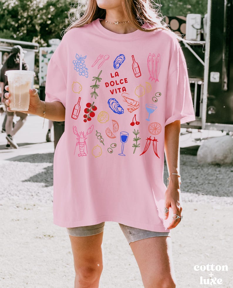 K&ouml;nnte beinhalten: Hellrosa Oversize-T-Shirt mit farbigen Illustrationen von Speisen und Getr&auml;nken, darunter Trauben, Austern und Flaschen. Der Schriftzug "LA DOLCE VITA" ist auf der Vorderseite aufgedruckt. Das Shirt ist aus weichem Material.
