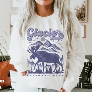 May include: White sweatshirt with a purple graphic design of a moose standing in front of mountains. The text "Glacier 1910 National Park" is printed below the graphic.