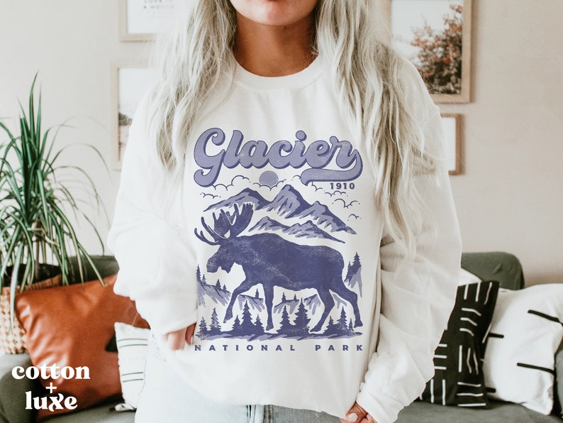 May include: White sweatshirt with a purple graphic design of a moose standing in front of mountains. The text "Glacier 1910 National Park" is printed below the graphic.