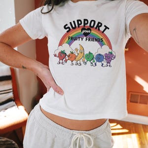 May include: White t-shirt featuring the slogan "SUPPORT your FRUITY FRIENDS" above a rainbow and cartoon fruit characters. The shirt is worn with light grey sweatpants. The words "cotton + luxe" are at the bottom left.