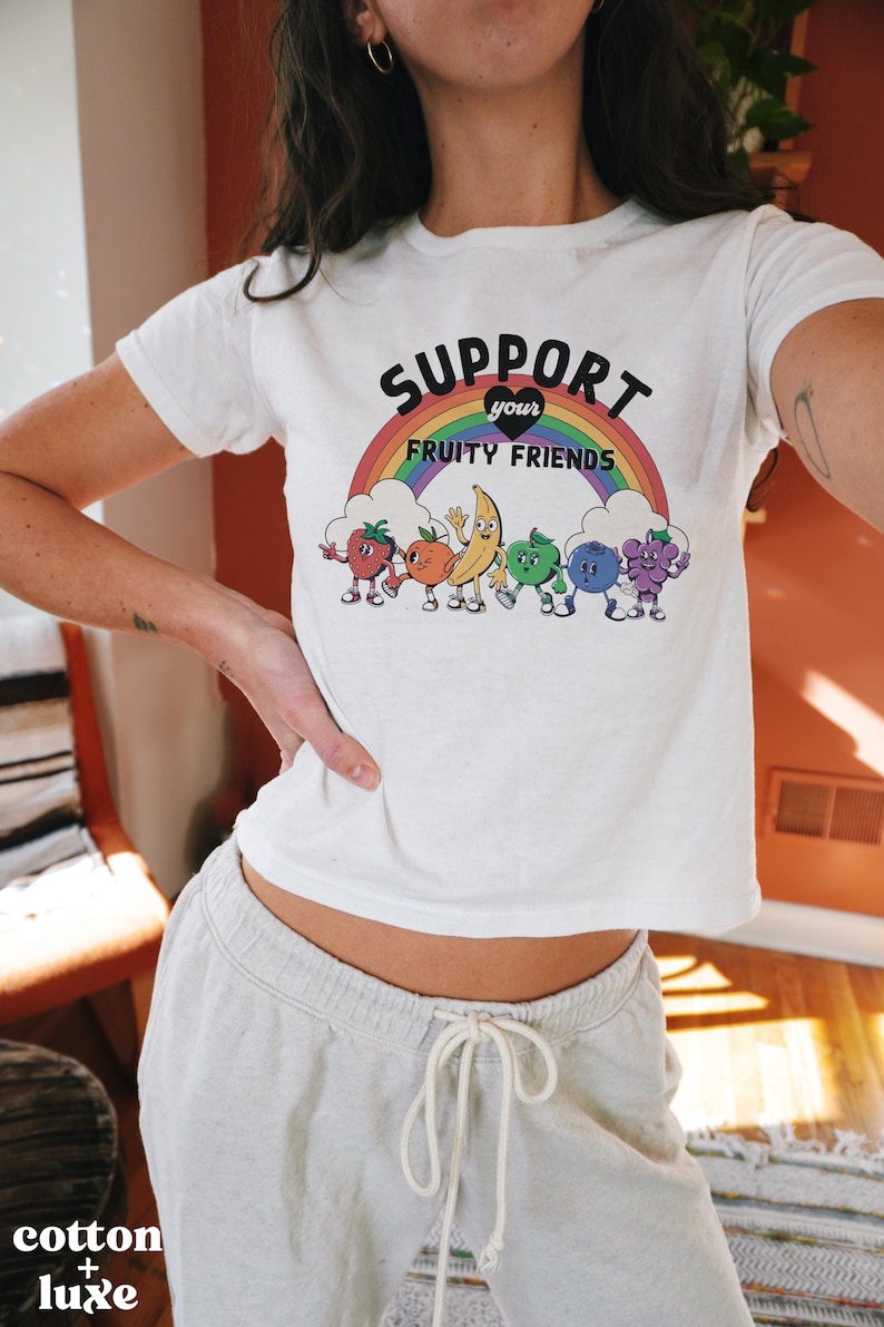 May include: White t-shirt featuring the slogan "SUPPORT your FRUITY FRIENDS" above a rainbow and cartoon fruit characters. The shirt is worn with light grey sweatpants. The words "cotton + luxe" are at the bottom left.
