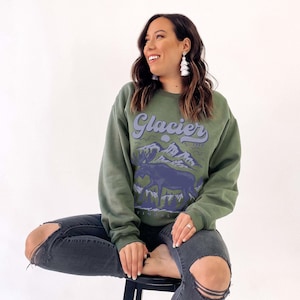 May include: A woman wearing a green sweatshirt with a blue and white graphic of a moose and mountains. The sweatshirt says "Glacier 1910" in a retro font.
