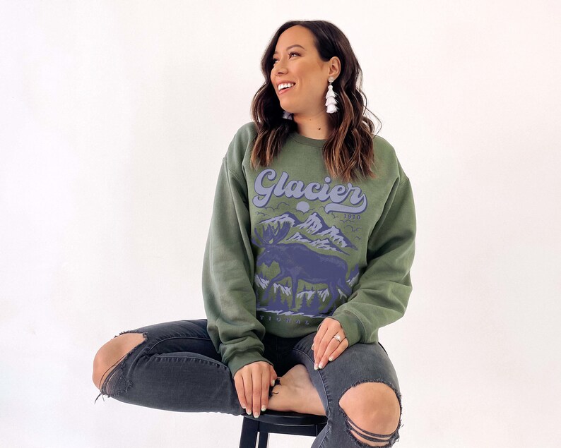 May include: A woman wearing a green sweatshirt with a blue and white graphic of a moose and mountains. The sweatshirt says "Glacier 1910" in a retro font.