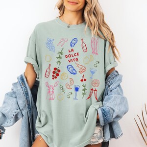 K&ouml;nnte beinhalten: Hellgr&uuml;nes T-Shirt mit einer farbenfrohen Illustration von Speisen und Getr&auml;nken, darunter Trauben, Austern und Weinflaschen. Der Text "LA DOLCE VITA" ist in Rot gedruckt. Das Shirt geh&ouml;rt zur cotton + luxe Kollektion.