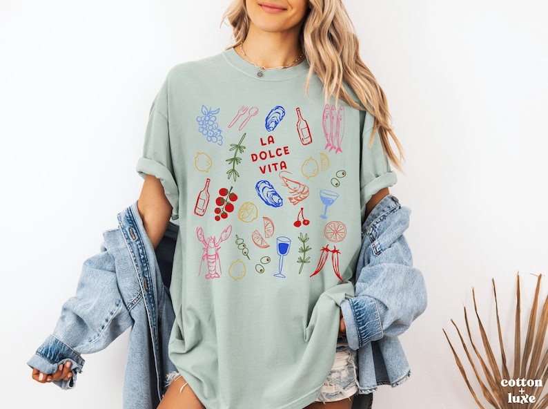 K&ouml;nnte beinhalten: Hellgr&uuml;nes T-Shirt mit einer farbenfrohen Illustration von Speisen und Getr&auml;nken, darunter Trauben, Austern und Weinflaschen. Der Text "LA DOLCE VITA" ist in Rot gedruckt. Das Shirt geh&ouml;rt zur cotton + luxe Kollektion.