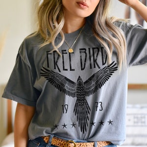 Puede incluir: Una camiseta gris con un gr&aacute;fico negro de un p&aacute;jaro con las alas extendidas y el texto "FREE BIRD" encima. El gr&aacute;fico tambi&eacute;n incluye los n&uacute;meros "19" y "73" y cinco estrellas.