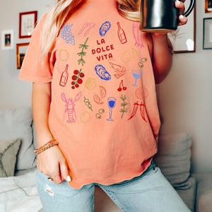 K&ouml;nnte beinhalten: Terracottafarbenes T-Shirt mit dem Schriftzug "LA DOLCE VITA" und farbenfrohen Illustrationen von Speisen und Getr&auml;nken. Das Design umfasst Trauben, Austern, Weinflaschen und einen Hummer. Das Shirt hat eine lockere Passform.