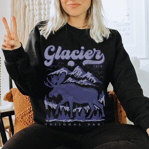 May include: Black sweatshirt with a purple graphic of a moose in front of mountains and the text "Glacier National Park 1910".