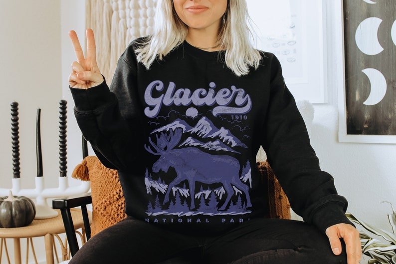 May include: Black sweatshirt with a purple graphic of a moose in front of mountains and the text "Glacier National Park 1910".