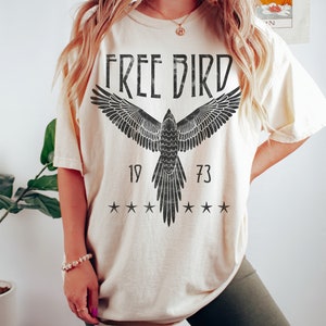 Puede incluir: Una camiseta de color crema con un gr&aacute;fico negro de un p&aacute;jaro con las alas extendidas. El gr&aacute;fico incluye el texto "FREE BIRD" y los n&uacute;meros "19" y "73".